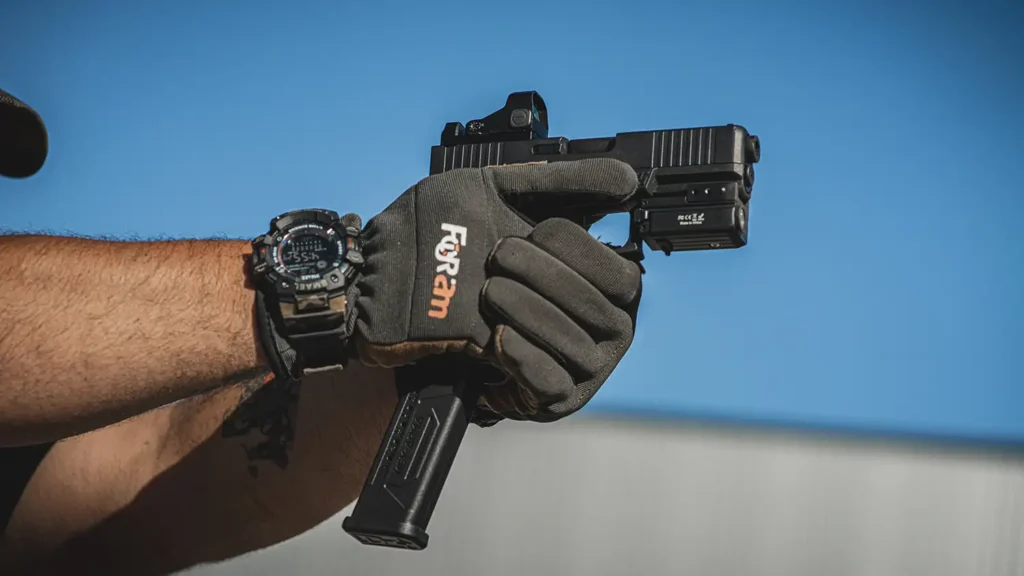 A gloved hand holds a black semi-automatic pistol with a red dot sight and weapon light.